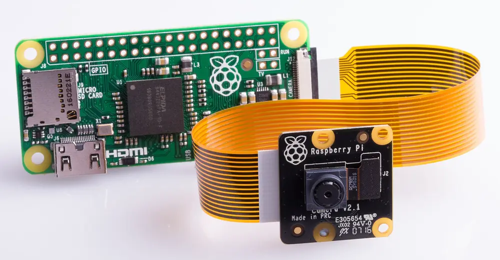 A Raspberry Pi Zero with a camera attached by a flat orange ribbon cable plugged into the connector on its short edge.